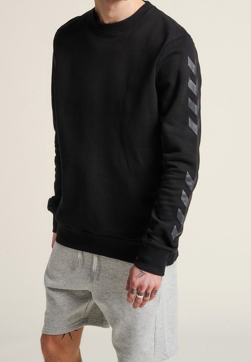 Personne portant un sweat-shirt noir avec des bandes chevrons sombres sur la manche et un short gris clair à cordon, debout devant un fond uni.