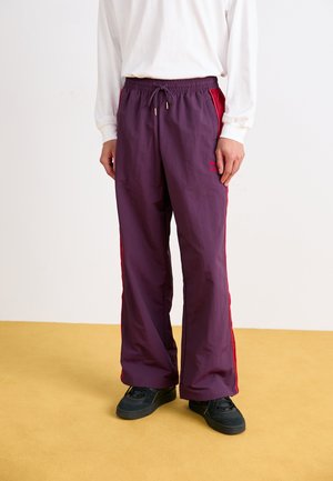 Purple track pants with red side stripes, elastic waistband, and drawstring. Made of lightweight fabric with a loose fit. Black sneakers.