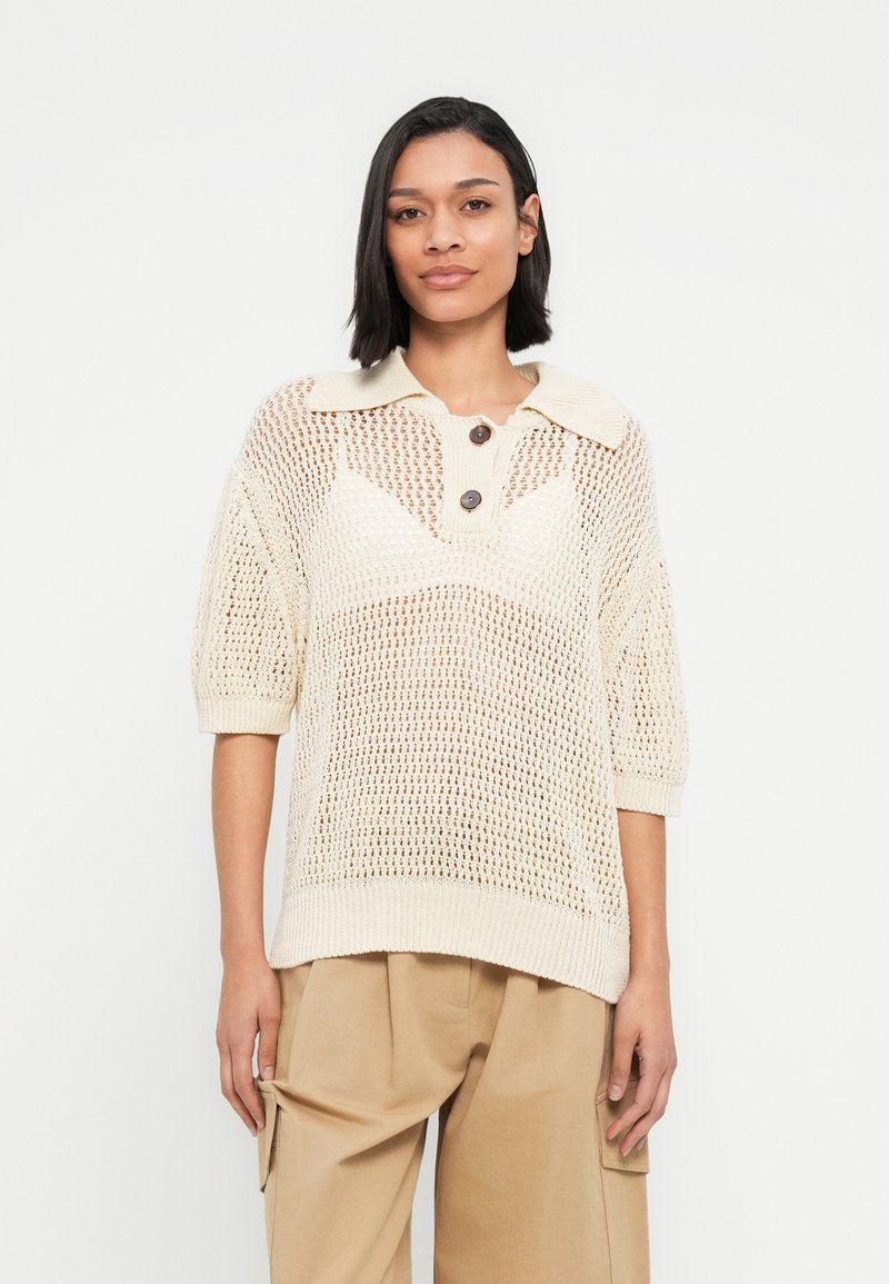 Pull-over tricoté couleur crème à coupe ample, manches courtes, col avec rabat et trois boutons marron ornés d'un motif texturé.