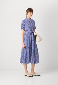 Vestido azul com padrão, mangas curtas, cintura ajustada e frente com botões. Combinado com sapatos planos em tom creme e uma bolsa branca.