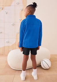 Blå fleecepullover med lange ærmer og høj krave. Kombineret med sorte shorts og hvide sportssko. Har en glat tekstur.