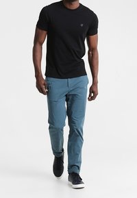 Camiseta negra de manga corta con un pequeño logo, combinada con pantalones ajustados en tono teal y zapatillas oscuras, caminando sobre un fondo neutro.