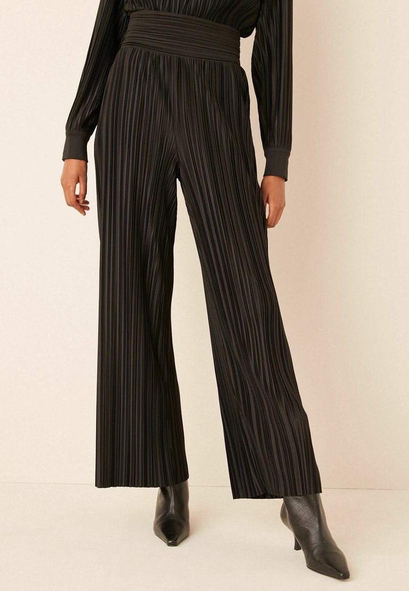 Femme portant un pantalon noir plissé, taille haute, à jambes larges, associé à des bottines noires à bout pointu sur un fond clair.