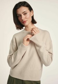 Beżowy sweter dziany z prążkowanymi mankietami i dołem, charakteryzujący się luźnym krojem i miękką fakturą. Szczegóły obejmują małe metalowe logo.