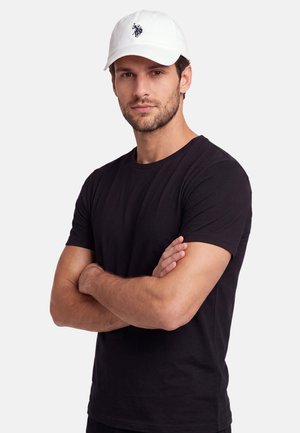 Man with beard wearing white cap and black t-shirt stands with arms crossed against plain background.