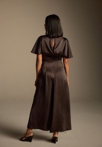 Lang, mørkebrun satin kjole med korte flagrende ærmer, en nøglehul detalje i ryggen og et flydende skørt. Båret med sorte kilehælsandaler.