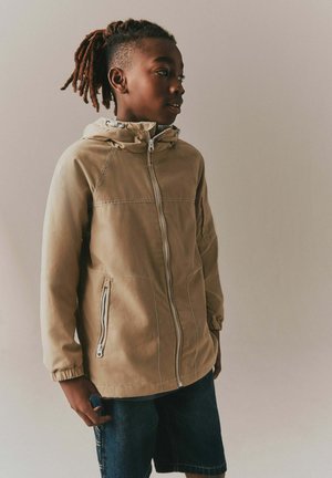 Dreng med fletninger iført en brun lynlåstrøje og denimshorts, stående sidelæns mod en ensfarvet beige baggrund.