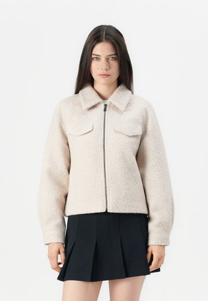 Femme aux longs cheveux noirs portant une veste zippée crème à texture avec col et une mini-jupe plissée noire, faisant face à l'avant sur fond blanc.