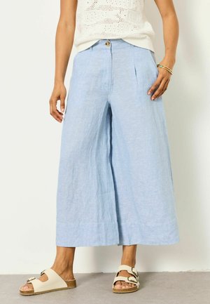 Frau trägt hellblaue, weit geschnittene, gekürzte Hosen, ein weißes ärmelloses Oberteil, beige Sandalen mit Schnallen und ein goldenes Armband mit türkisen Steinen.