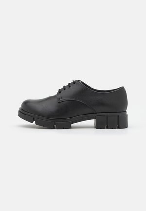 Chaussure Oxford en cuir noir à lacets, semelle en caoutchouc épaisse et segmentée avec un petit talon sur fond blanc uni.