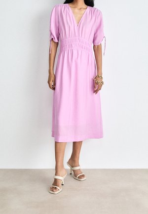 Vrouw draagt een lichtroze midijurk met korte mouwen, V-hals en geplooide taille, gecombineerd met beige sandalen met hak en gouden armbanden.
