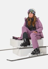Skidragt i lys lilla, med en jakke med sort mønsterlag, matchende bukser, sorte skistøvler og en hvid hjelm med spejlevisir.