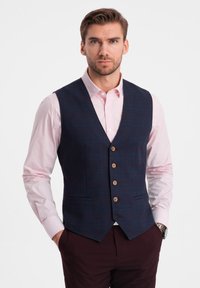 Gilet a quadrati blu con cinque bottoni in legno, orlo a punta e tasche laterali, indossato sopra una camicia a maniche lunghe rosa chiaro.