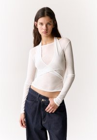 White long-sleeve top with a wrap design, sheer texture, and a square neckline. Paired with dark blue high-waisted denim jeans.