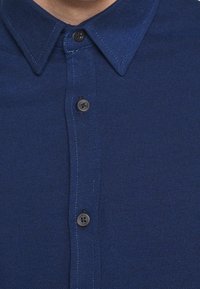 Camicia blu navy con bottoni, realizzata in tessuto strutturato, caratterizzata da colletto a punta, patta con bottoni neri e dettagli di cuciture a contrasto.