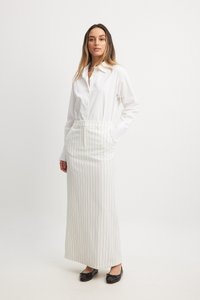Chemise blanche à boutons assortie d'une longue jupe blanche à rayures. La jupe possède une taille avec des poches et des chaussures plates noires.