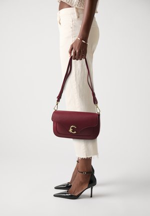 Woman wearing cream pants and black heels, holding a burgundy leather handbag with gold clasp by its strap.