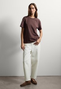 T-shirt in cotone marrone, vestibilità comoda, maniche corte; abbinato a jeans bianchi a gamba larga e scarpe basse marroni su uno sfondo neutro.