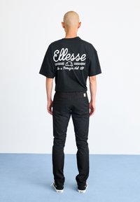 Fekete rövid ujjú póló fehér "Ellesse" logóval a hátsó részen, fekete nadrággal és fekete sportcipővel, világoskék háttér előtt.
