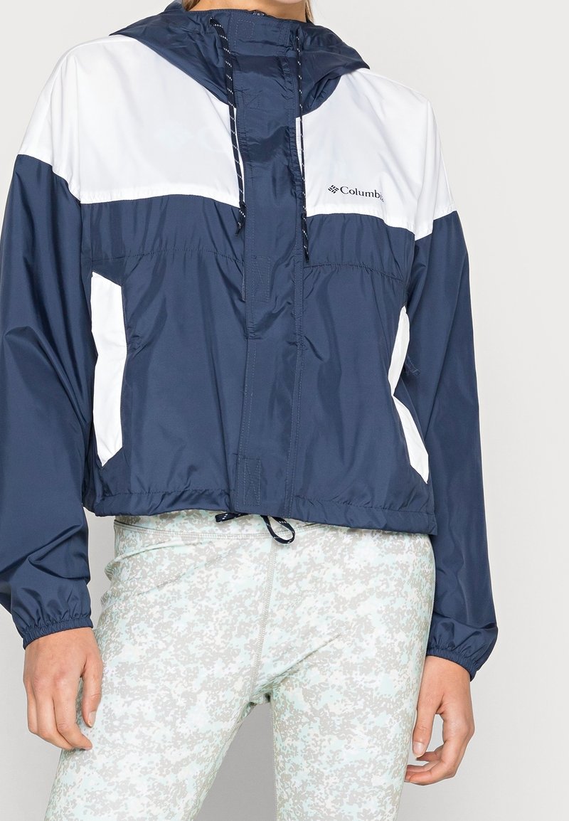 Granatowa kurtka wiatrówka z białymi akcentami, krótki fason, przednie kieszenie, kaptur ze ściągaczem oraz elastyczne mankiety. Lekki, wodoodporny materiał.