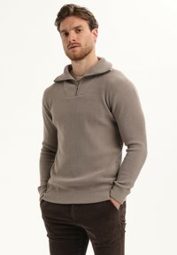 Lichtbruin zip-neksweater met een gestructureerde weving, voorzien van een kleine kraag en lange mouwen, gecombineerd met donkerbruine broek.