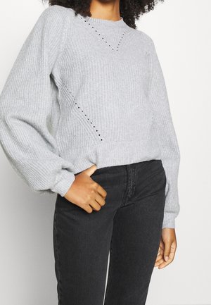 Personne portant un pull en maille gris clair avec des manches ballon et un jean noir taille haute, main dans la poche, debout devant un fond blanc.