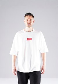 White oversized t-shirt with short sleeves, featuring a red logo patch on the chest. The fabric appears smooth and lightweight.