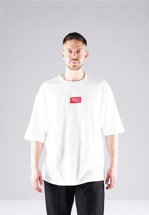 Weißes übergroßes T-Shirt mit kurzen Ärmeln, das auf der Brust mit einem roten Logo-Patch versehen ist. Der Stoff wirkt glatt und leicht.