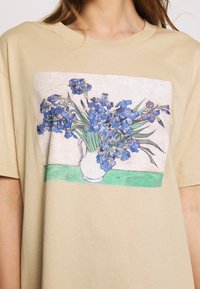 Beige bomull t-shirt med en rektangulär illustration av blå iris i en vit vas på en grön yta. Kort ärm och avslappnad passform.