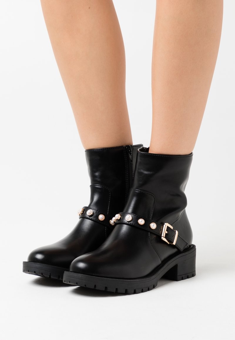Czarne skórzane botki za kostkę z zamkiem błyskawicznym, z przednim paskiem ozdobionym akcentami przypominającymi perły oraz masywnym obcasem z teksturowaną podeszwą.