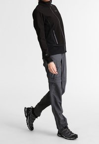 Chaqueta negra con cremallera y cuello alto, pantalones cargo grises y zapatillas deportivas negras con suelas texturizadas y diseños curvos.