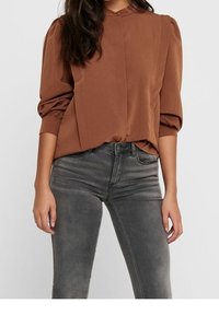 Braune Bluse mit hohem Kragen, puffigen Ärmeln und Vorderfalten, kombiniert mit grauen skinny Jeans in einem verwaschenen Finish.