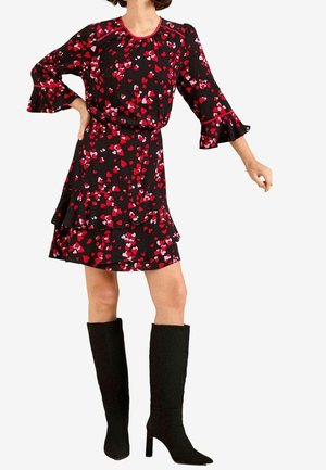 Kobieta w czarnej sukience z wzorem czerwonych i różowych serc, z falbanami na rękawach i krawędzi, w połączeniu z czarnymi, sięgającymi kolan, butami na obcasie.