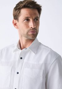 Weißes Kurzarmhemd mit strukturierter Oberfläche, ausgestattet mit zwei Brusttaschen, einem Button-Down-Kragen und dunklen Knöpfen.