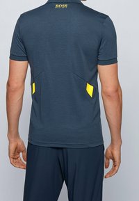 Hombre vestido con una camiseta polo azul marino de Hugo Boss con acentos amarillos, visto desde la espalda contra un fondo gris claro liso.