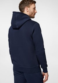 Granatowa bluza z kapturem wykonana z mieszanki bawełny, z regulowanym kapturem, ściągaczami przy mankietach i u dołu. Gładka faktura z minimalnymi detalami szwów.