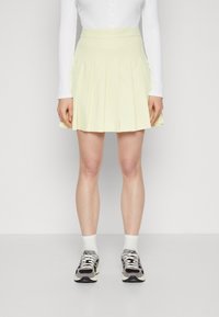 Plisovaná sukně nad kolena v měkké žluté tkanině, s přiléhavým pasem. Nosit se bílými ponožkami a vzorovanými černými teniskami.