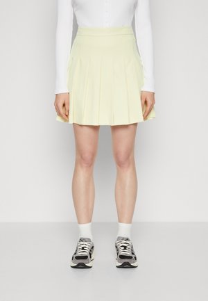 Jupe plissée au-dessus du genou en tissu jaune doux, dotée d'une taille ajustée. portée avec des chaussettes blanches et des baskets noires à motifs.