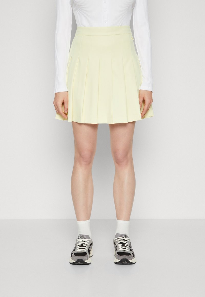 Plisovaná sukně nad kolena v měkké žluté tkanině, s přiléhavým pasem. Nosit se bílými ponožkami a vzorovanými černými teniskami.