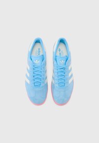 Des baskets en velours bleu présentent des rayures blanches, une semelle en caoutchouc rose et une languette bleu clair, avec un logo sur le côté et la semelle intérieure.