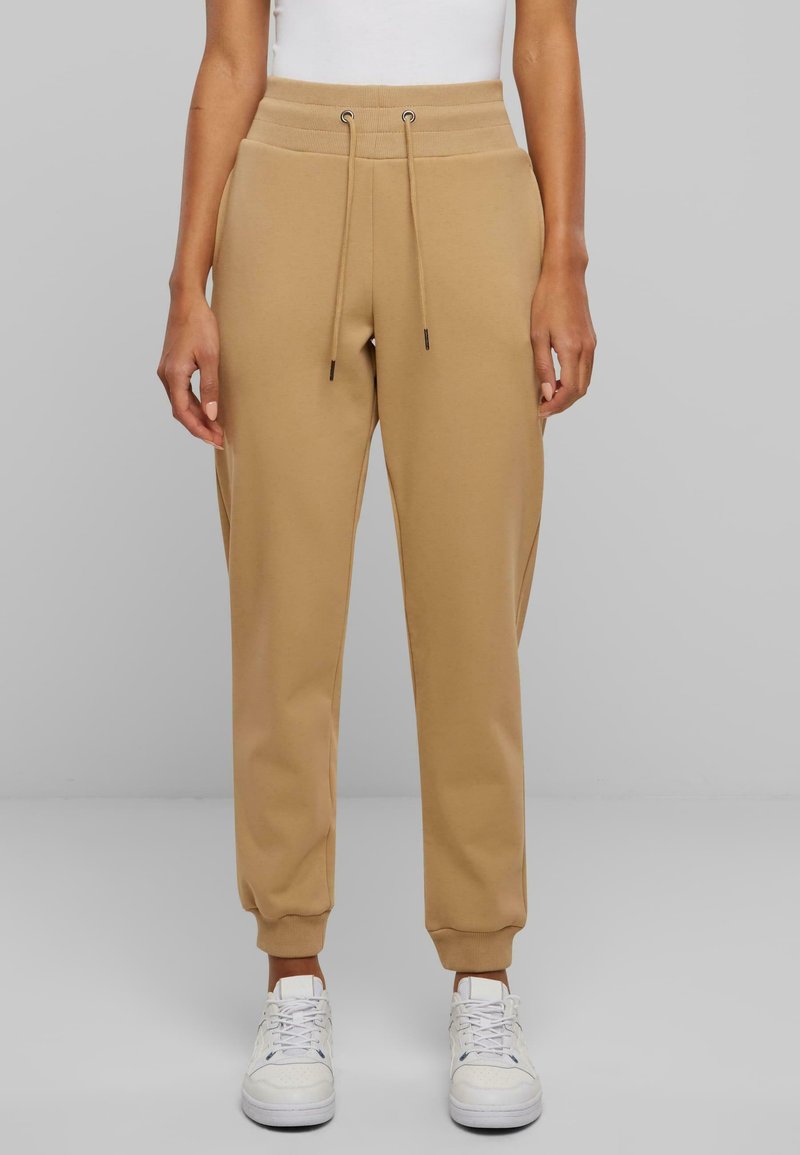 Beige joggebukser laget av myk, elastisk stoff med snor i midjen, avrundede ankler og sidelommer, paret med hvite sneakers.