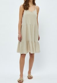 Kvinna klädd i en ärmlös, beige, lager-på-lager midi-klänning med tunna remmar och platta, flätade sandaler mot en neutral bakgrund.