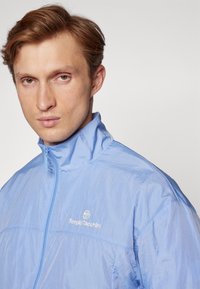 Lyseblå zip-up jakke med høj krave, der har en glat, let tekstur og et hvidt logo på venstre bryst.