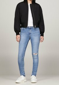 Mujer vestida con jeans skinny rasgados de color azul claro, zapatillas blancas, camiseta blanca y una chaqueta bomber recortada negra, de pie contra un fondo liso.