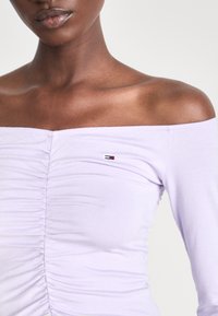 Fioletowa bluzka typu off-the-shoulder z marszczonym materiałem i małym logo na piersi. Wykonana z elastycznej tkaniny, ma długie rękawy.