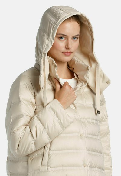 Beige pufferjack met een hoge kraag en een afneembare capuchon. Het heeft gewatteerde secties, drukknoopsluitingen en zijzakken. Gladde, glanzende textuur.