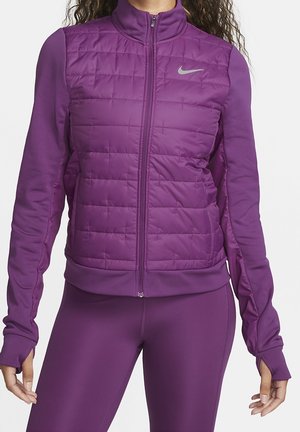 Frau trägt eine lila gesteppte ärmellose Jacke über einem langärmeligen Oberteil und passenden enganliegenden Leggings.