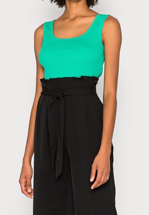 Femme portant un haut turquoise sans manches rentré dans un pantalon noir taille haute avec une ceinture en tissu nouée à la taille.