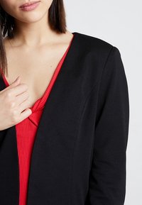 Černý texturovaný blazer přes červený ribbovaný top s hlubokým výstřihem do V. Blazer má přiléhavý střih, což ukazuje na vrstvený design.