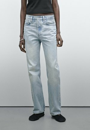 Personne portant un jean droit bleu clair et un top court noir, debout devant un fond gris clair uni.